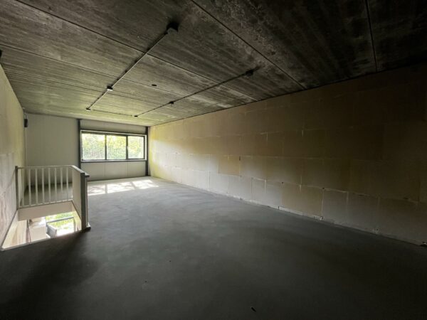 Lege ruimte met betonnen vloer en plafond, groot raam aan het einde en trapgat met witte leuning links.